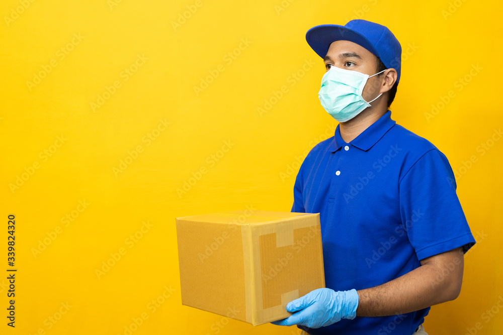 Delivery man blue uniform wearing rubber gloves and mask holding parcel cardboard box on yellow background. Delivery man blue uniform wearing rubber gloves and mask holding parcel cardboard box on yellow background.