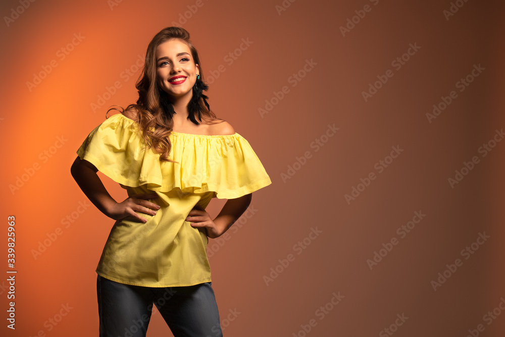 Obraz premium Studio portrait of young beautiful woman with long curly hair and perfect make up wearing a yellow blouse and red earrings on orange background. Text space.