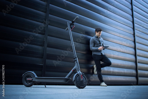 Electric scooter. Eco friendly modern way of transportation. In background handsome business person caucasian man using tablet computer leaning on black business building.