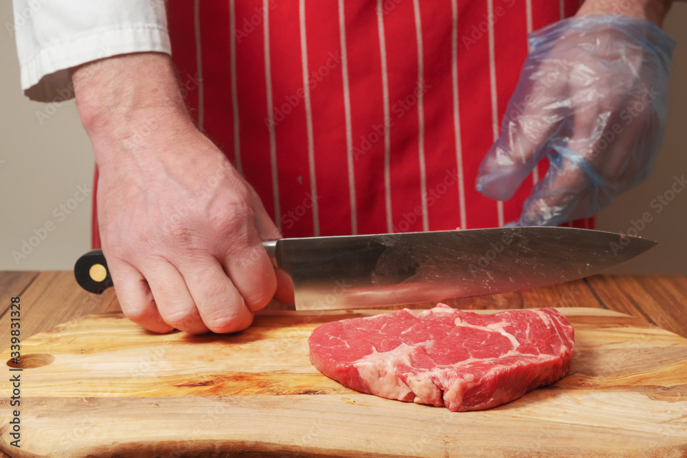 Rib eye steak on wooden cutting board, professional butcher with knife in his hand, Butcher dressed in white coat, red apron and blue glove.