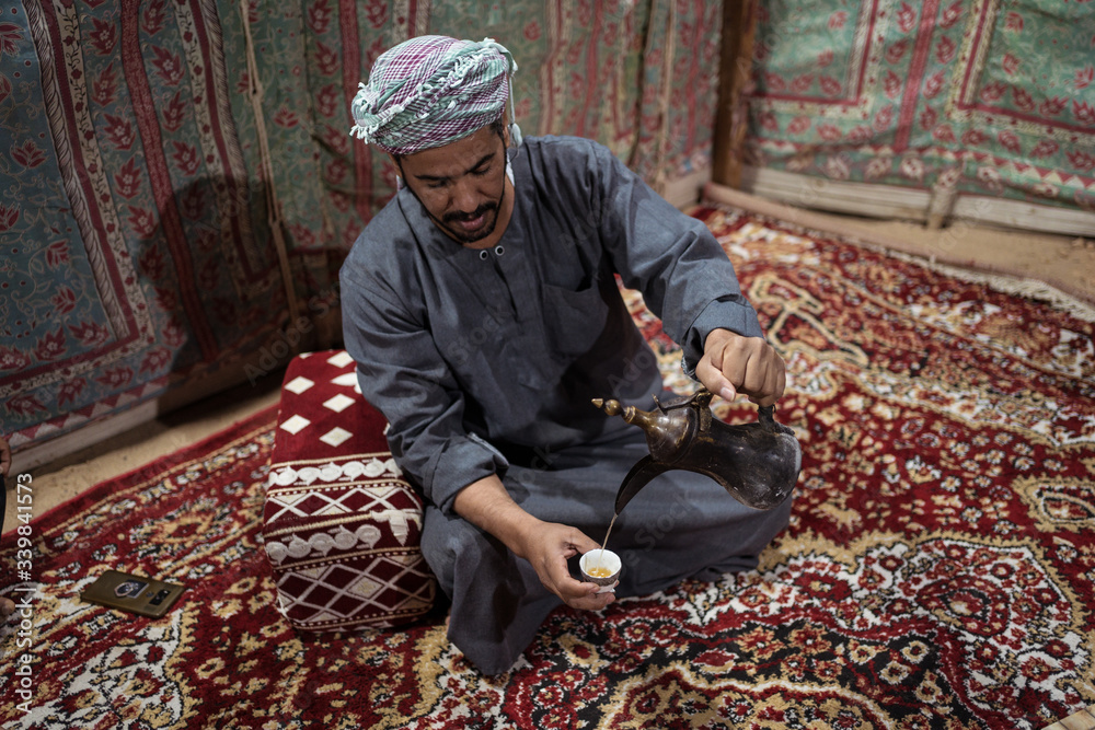 Bedouin man wearing traditional clothes prepares tea on a carpet in the Saudi desert