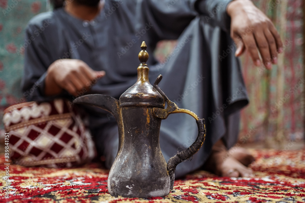 Closeup of traditional Arab teapot with Bedouin man wearing traditional clothes sitting on a carpet in the Saudi desert