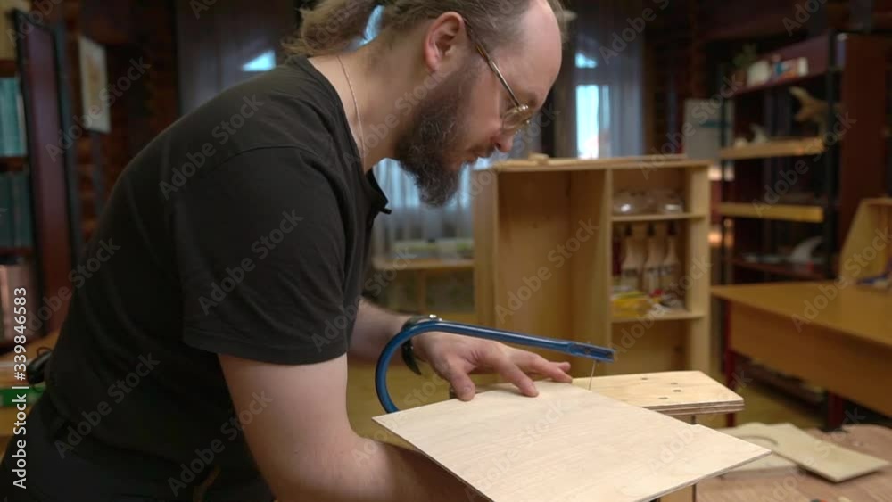 Sunday school teacher with long hair in ponytail and beard cuts brown plywood on support with blue jigsaw in classroom Sunday school teacher with long hair in ponytail and beard cuts brown plywood on support with blue jigsaw in classroom