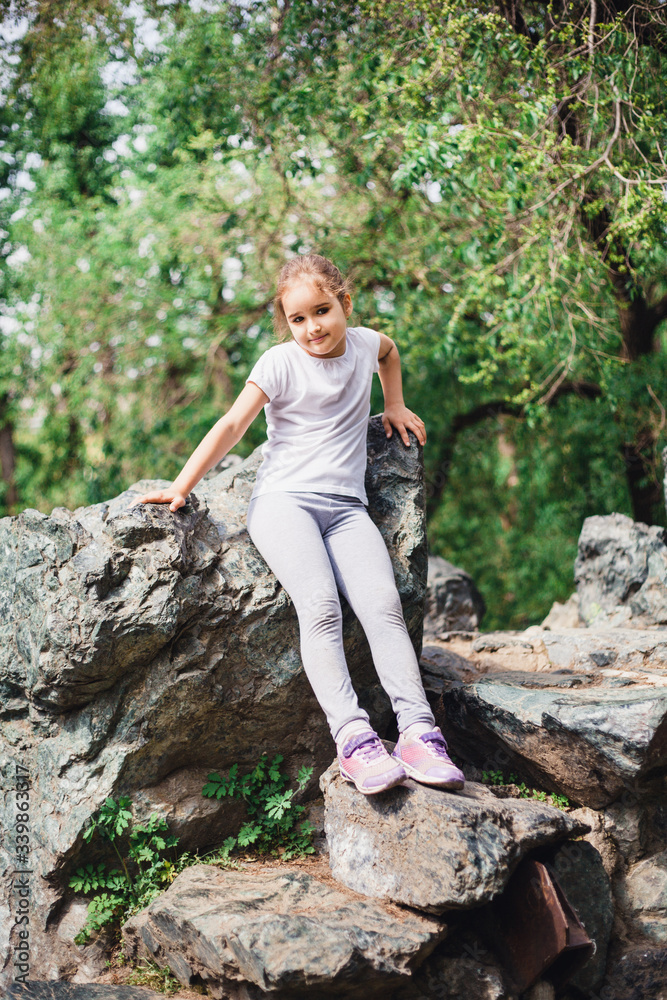 Naklejka premium A little girl in a t-shirt and pants climbs on rocks, mountains, peaks, conquering peaks, game, Park, forest, adventure with family, learning geography, terrain