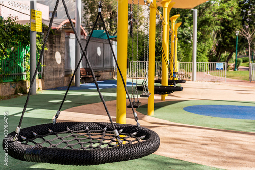 Black round mesh swing, empty yellow playground. No children, parents, no people. Sunny weather, park, warm. Virus and quarantine. The concept of protecting and preventing viruses and epidemics. Urban