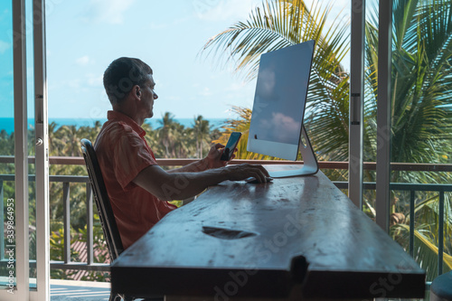 Man works at home at a wooden table with a laptop and smartphone with sea and palm trees in the background
 photo