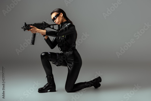 futuristic african american woman in glasses aiming assault rifle while standing on knee on grey