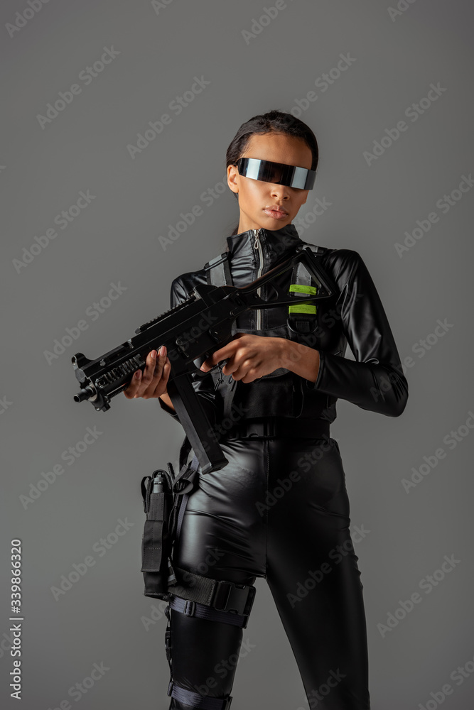 attractive futuristic african american woman in glasses with assault rifle isolated on grey attractive futuristic african american woman in glasses with assault rifle isolated on grey