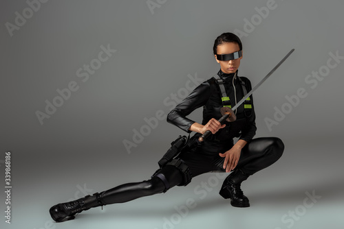 attractive futuristic african american woman in glasses sitting with sword on grey
