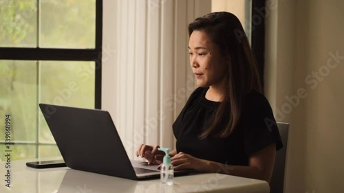 Serious asian lady working on laptop. Hand gel sanitizer on the table to prevent spread of coronavirus. Working from home and social distancing.