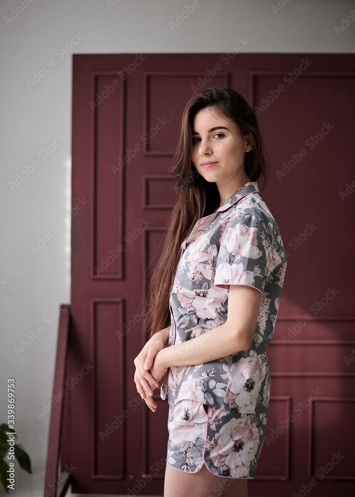 Young pretty slim brunette woman, wearing colorful light pink and grey pajamas with flowers pattern, posing for picture in front of claret marsal wall. Sleepwear design. Cute girl in the morning. Young pretty slim brunette woman, wearing colorful light pink and grey pajamas with flowers pattern, posing for picture in front of claret marsal wall. Sleepwear design. Cute girl in the morning.