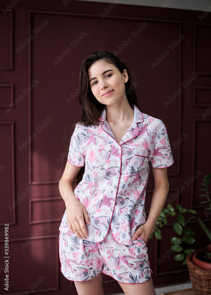 Young pretty slim brunette woman, wearing colorful light pink and grey pajamas with flowers pattern, posing for picture in front of claret marsal wall. Sleepwear design. Cute girl in the morning. Young pretty slim brunette woman, wearing colorful light pink and grey pajamas with flowers pattern, posing for picture in front of claret marsal wall. Sleepwear design. Cute girl in the morning.