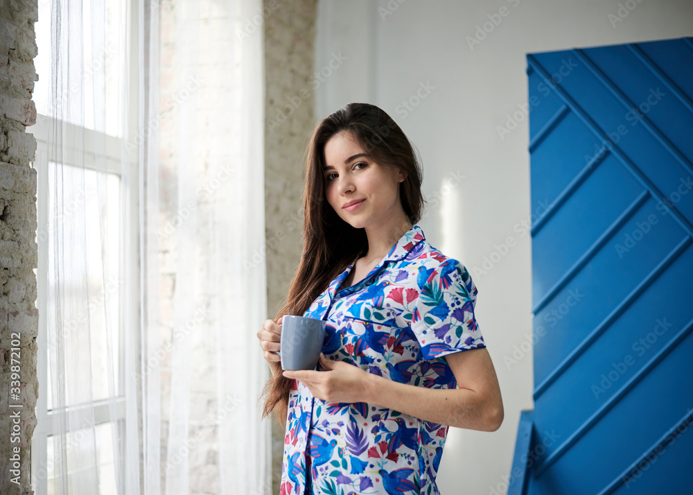 Young pretty slim brunette woman, wearing colorful blue pajamas with flowers pattern, holding grey coffee cup mug by the window with white tulle. Sleepwear design. Cute girl, drinking morning tea. Young pretty slim brunette woman, wearing colorful blue pajamas with flowers pattern, holding grey coffee cup mug by the window with white tulle. Sleepwear design. Cute girl, drinking morning tea.
