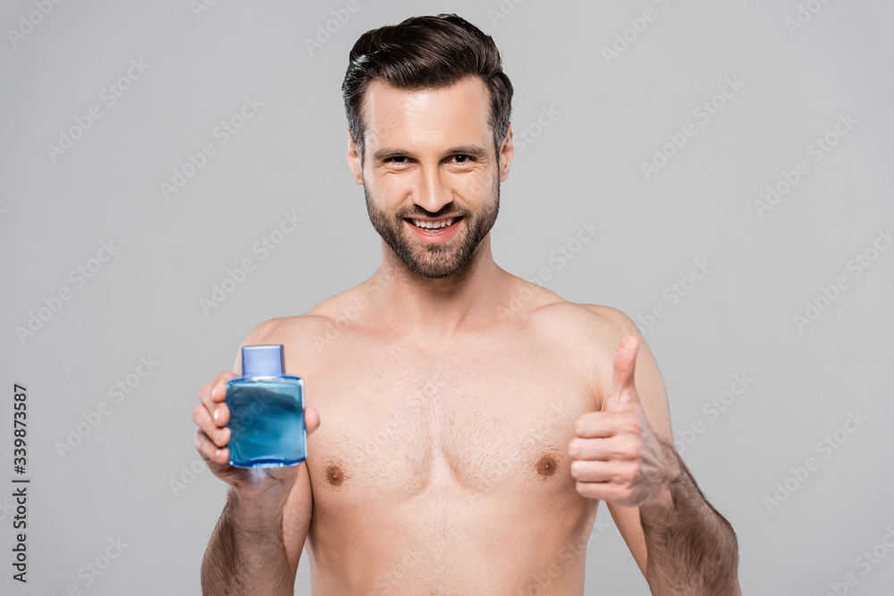 happy man holding bottle with after shave lotion and showing thumb up isolated on grey happy man holding bottle with after shave lotion and showing thumb up isolated on grey