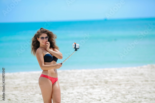 Woman takes a selfie in the background of the ocean. The girl in the swimsuit is young and beautiful with a selfie stick in her hands posing and photographed