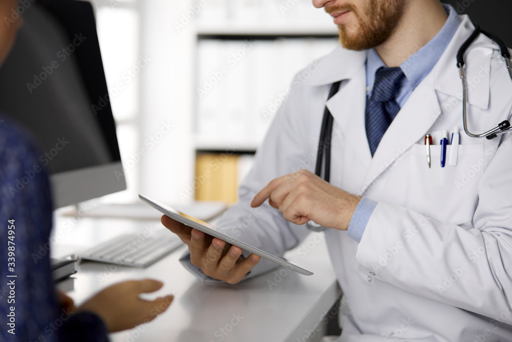 Fototapeta premium Unknown red-bearded doctor and patient woman discussing current health examination while sitting and using tablet computer in clinic, close-up. Medicine concept