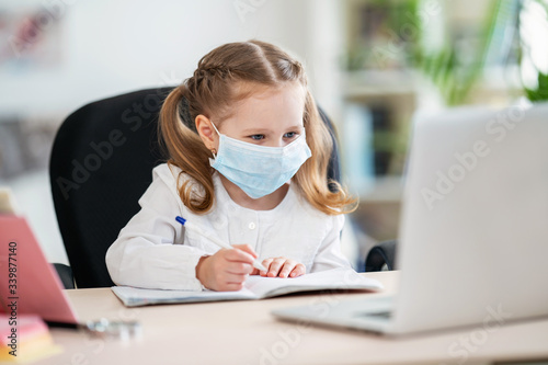 Cute little girl, doing homework, writing in notebook, using laptop, e-learning