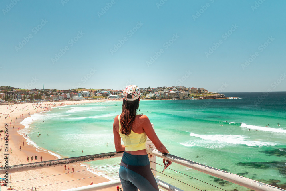 Naklejka premium Woman at Bondi Beach, Sydney, Australia. Girl in work out gear looking at view of the ocean, sun, sea and sand scene, while on vacation. Holiday, tropical, fitness concepts. 