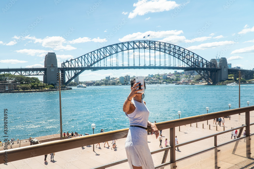 Fototapeta premium Woman taking selfie with mobile phone at iconic Sydney Harbor Bridge. Cityscape, water, with buildings in CBD area. Tourist attraction in Australia. Technology, tourism, travel, taking a photo concept