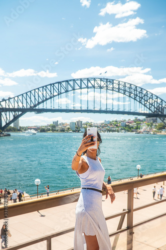 Woman taking selfie with mobile phone at iconic Sydney Harbor Bridge. Cityscape, water, with buildings in CBD area. Tourist attraction in Australia. Technology, tourism, travel, taking a photo concept