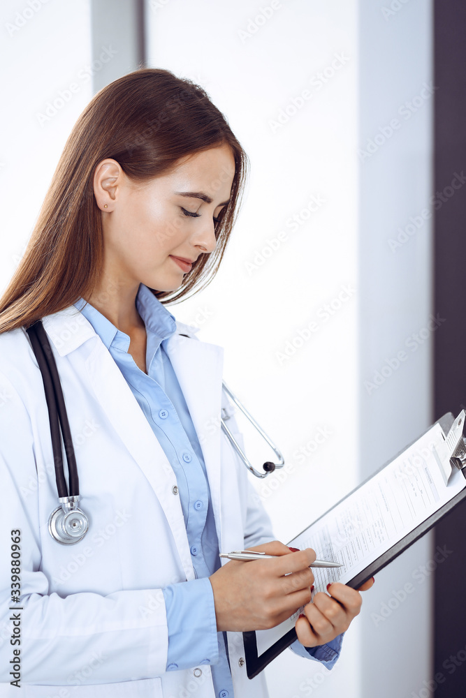 Doctor woman filling up medical form while standing near window in clinic. Physician at work. Medicine and health care concept Doctor woman filling up medical form while standing near window in clinic. Physician at work. Medicine and health care concept