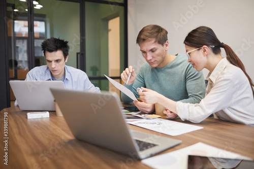 Businessman and businesswoman sitting at the table with laptop and discussing document with their colleague sitting near by and using his laptop at office