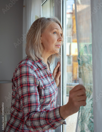 An aged woman is standing in a room by the window, smiling. © Albina