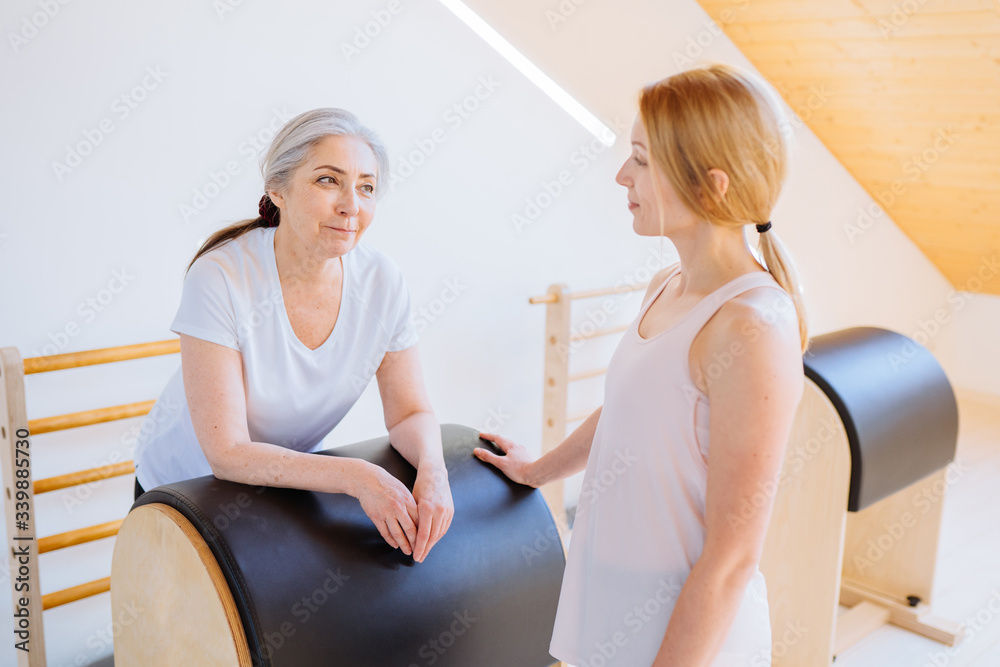 Young professional instructor helping an elderly woman can't workout independently doing stretching back, relaxing muscles exercise on pilates barrel equipment in studio. Young professional instructor helping an elderly woman can't workout independently doing stretching back, relaxing muscles exercise on pilates barrel equipment in studio.