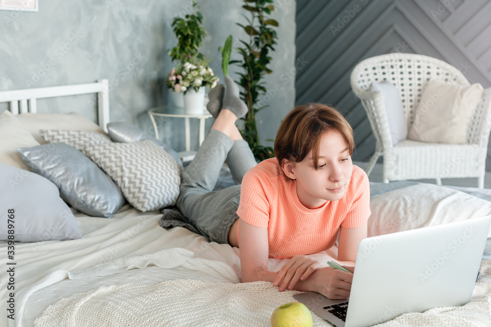 A teenage girl at home is lying on a bed with a laptop in headphones.