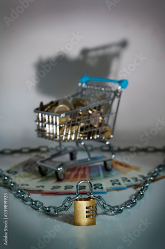 Small shopping cart with Money in Euro coins and banknotes. Dark and ominous setting, spooky. Chain with safety lock for protection against thieves and criminals. 