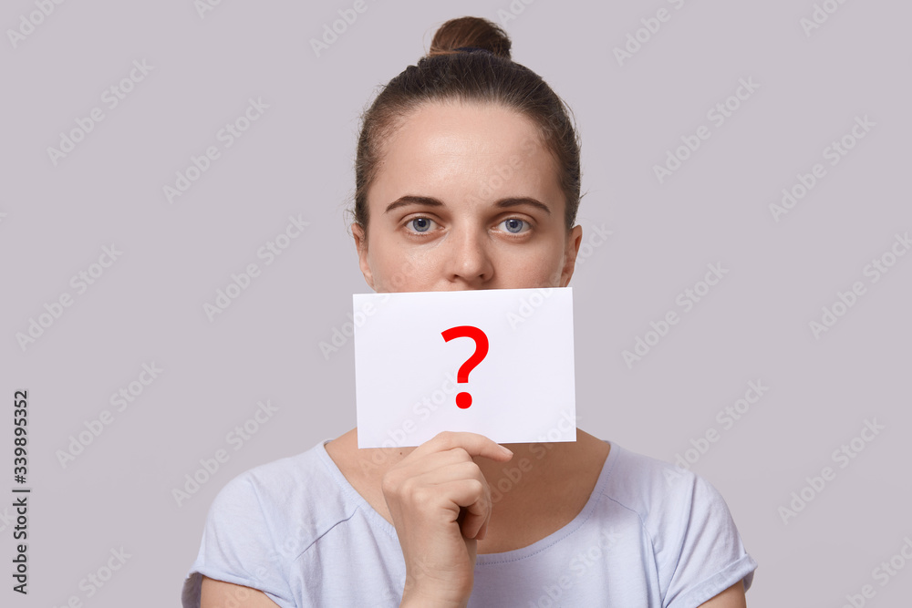 Closeup portrait of winsome lady with white card near lips, has red question mark on sheet of paper, woman with hair bun wearing casual white t shirt, girl looking directly at camera. People concept. Closeup portrait of winsome lady with white card near lips, has red question mark on sheet of paper, woman with hair bun wearing casual white t shirt, girl looking directly at camera. People concept.