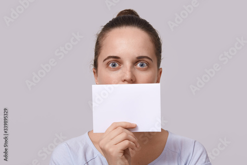 Studio shot of shocked woman with hair bun, holding empty card near her lips, posing with big eyes, covers her mouth with piece of paper with copy space for inscription, isolated on studio background.