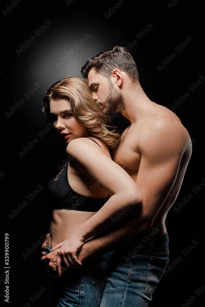 Naklejka premium sexy muscular man embracing and kissing attractive girl in bra on black background