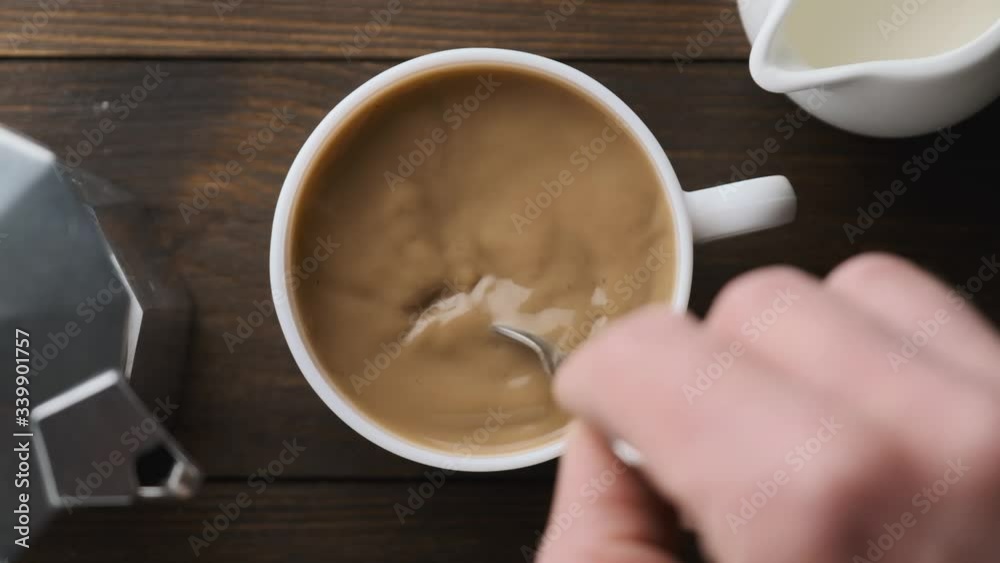 Male hand stirring coffee and milk with spoon on wooden table, top view, slow motion