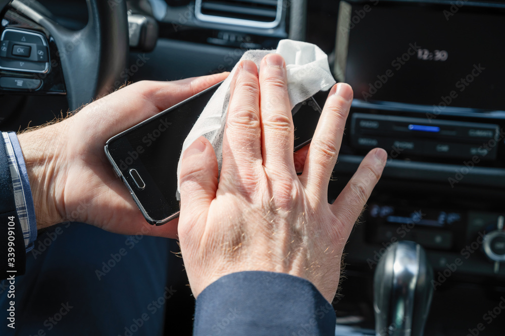 Middle age businessman in a blue suit cleaning mobile phone or gadget in a car using antivirus antibacterial wet wipe (napkin) for protect himself from bacteria and virus.