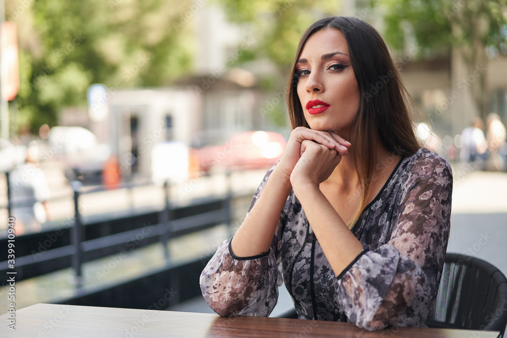 Beautiful elegant young girl with dark dyed long hair sits at a table summer outdoor cafe waiting for her boyfriend.