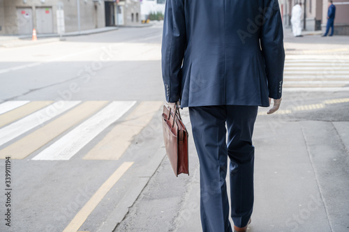 Alone businessman in a blue suit with briefcase in a protective white rubber gloves on the crosswalk on the city street. Health protection against viruses, germs and bacteria. Coronavirus