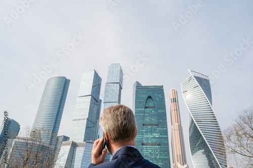 Alone middle-aged gray-haired businessman in a blue suit calling by phone standing opposite Moscow City skyscrapers.Low angle view. Russia