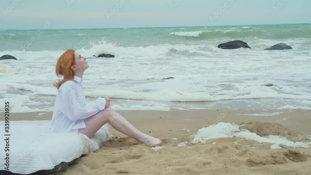 lonely, saddest girl sitting on the shore of the cold sea in bed. winter wind autumn or spring cold weather nude woman in white shirt. frozen. white bed. ocean. against the background of big waves lonely, saddest girl sitting on the shore of the cold sea in bed. winter wind autumn or spring cold weather nude woman in white shirt. frozen. white bed. ocean. against the background of big waves
