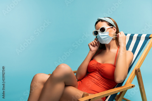 Woman in sunglasses putting medical mask on face on deckchair isolated on blue