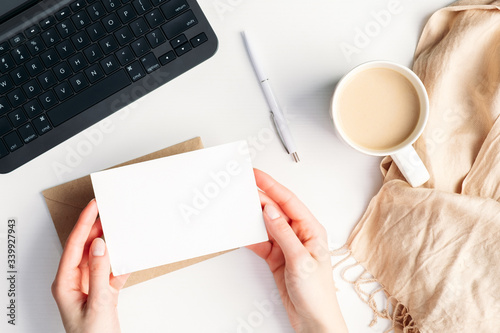 Female hands holding blank invitation card mockup over cozy home desk with cup of coffee, blanket, laptop computer. Hygge, autumn fall, comfort concept