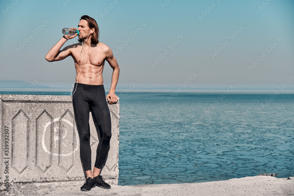 Shot of Topless Healthy Handsome Active Man With Fit Muscular Body standing outdoors holding water bottle. Fitness man taking a break after running workout.