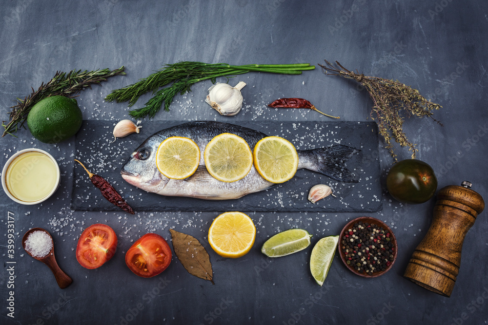 Dorado fish lies on a black stone cutting Board on beautiful gray concrete. Next are: lime, lemon, tomato, pepper, garlic, spices, salt and herbs Dorado fish lies on a black stone cutting Board on beautiful gray concrete. Next are: lime, lemon, tomato, pepper, garlic, spices, salt and herbs