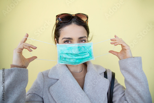 Donna  bianca con i capelli castani infila una  una mascherina chirurgica e guarda seria, isolata su sfondo giallo  photo