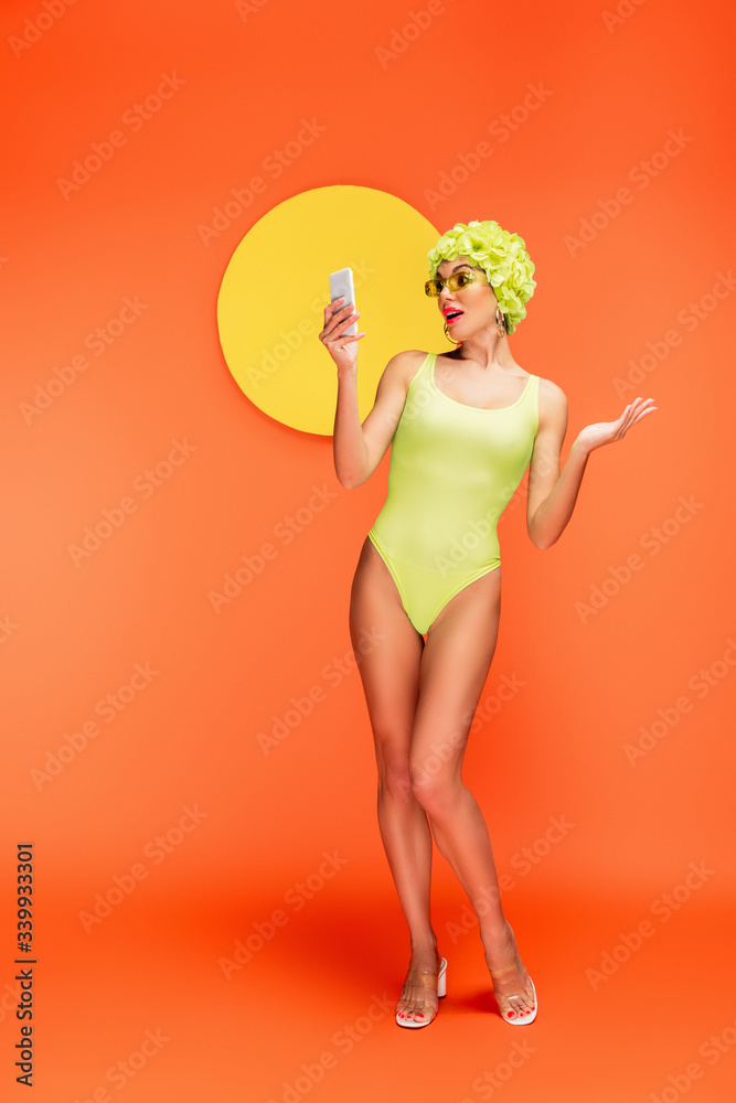 Shocked woman in hat with decorative flowers looking at smartphone with yellow circle behind on orange Shocked woman in hat with decorative flowers looking at smartphone with yellow circle behind on orange