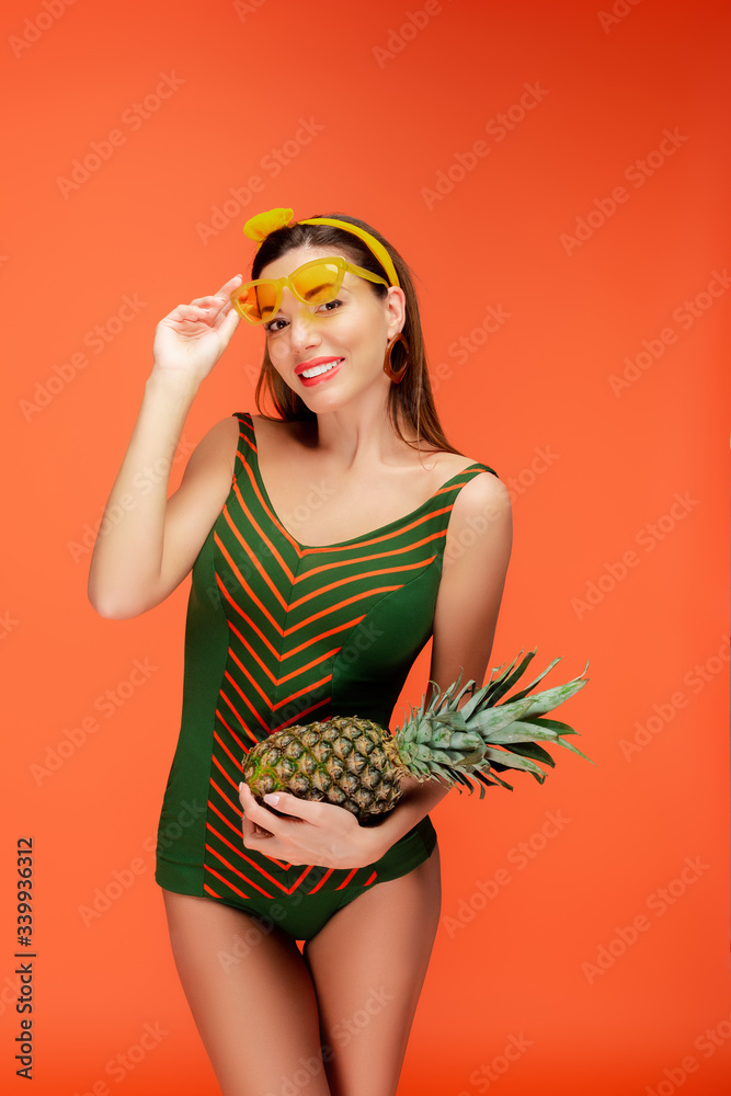 Front view of woman with sunglasses and pineapple smiling and looking at camera isolated on orange Front view of woman with sunglasses and pineapple smiling and looking at camera isolated on orange