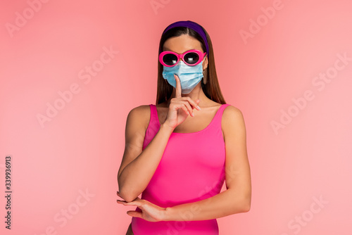 Front view of woman in sunglasses and medical mask showing shh sign isolated on pink