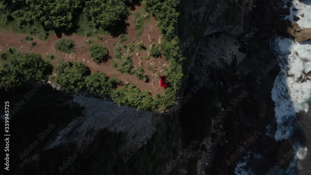 Aerial drone view of happy fit hipster young woman in red dress freestyle dance outdoors standing on cliff edge with amazing ocean and cloudy sky view. Adventure lifestyle youth and happiness concept. Aerial drone view of happy fit hipster young woman in red dress freestyle dance outdoors standing on cliff edge with amazing ocean and cloudy sky view. Adventure lifestyle youth and happiness concept.