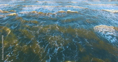 Aerial dynamic view to green sea illuminated by sun moving towards together with breaking waves with sheets of white spume and mystical shadows on the water till they meet seashore with golden sand