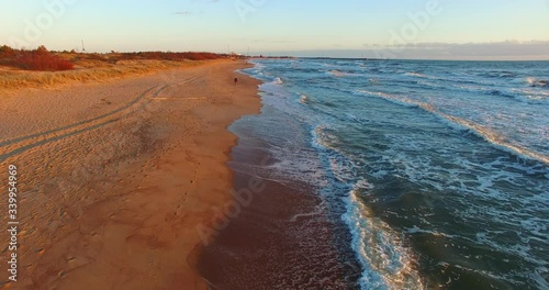 Aerial dynamic view from a top following from behind a woman walking along the sea, leaving imprints on wet sand next to the water, avoiding huge foamy waves of blue sea